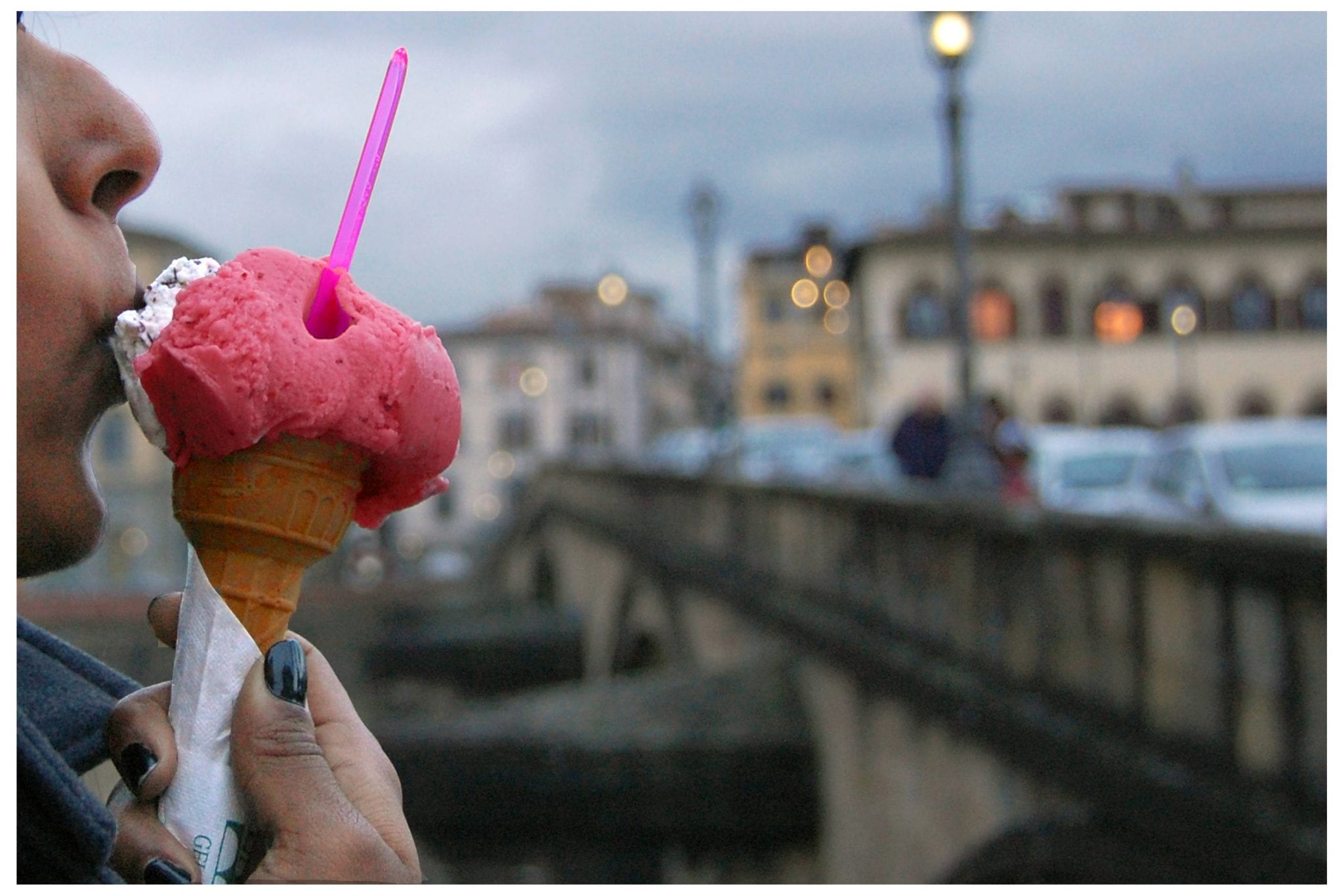 a close up of a woman eating an icecream or gelato, with the blurred skyline of an italian city, showing a bridge with lamposts and pastel coloured buildings in the background