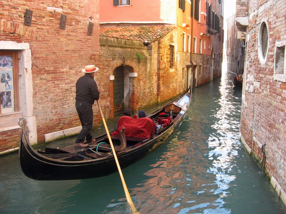 A couple and a gondolier wearing a straw hat, glide on a gondola down a narrow canal of Venice, with brick buildings with windows on wither side.