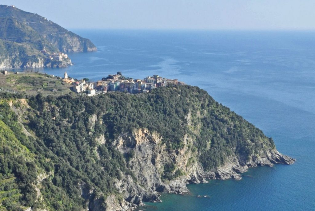 A rugged cliff side with small village on the top in a coastal area of italy called the Cinque Terre, with another cove behind it and a vast blue sea to the right