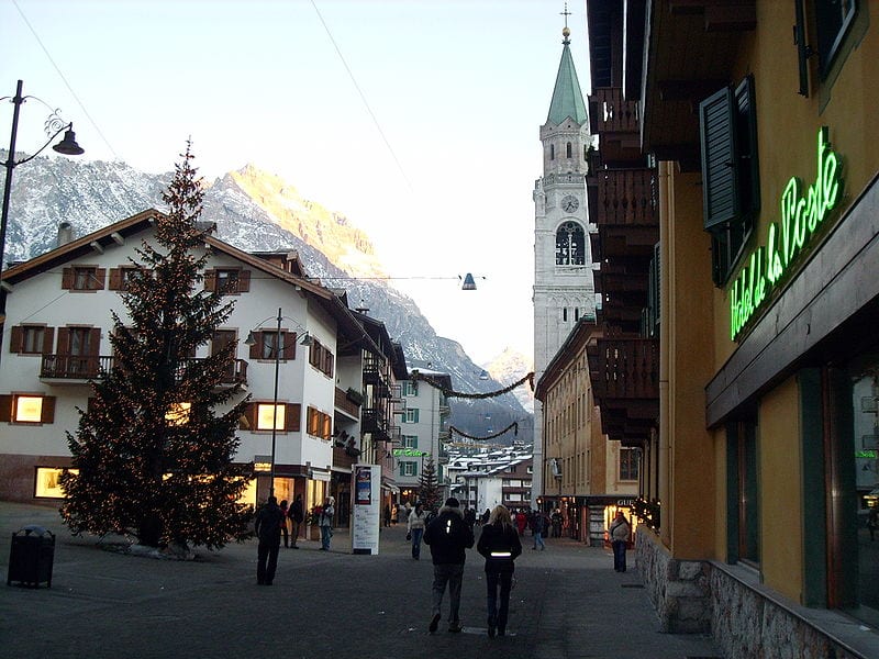 For one of the best towns in the doloites to visit to ski go to Cortina d'Ampezzo. A street in the town with a large fir tree next to the traditional chalet style buildings and the church spire on the opposite side of the street, with the snowy mountains behind. 