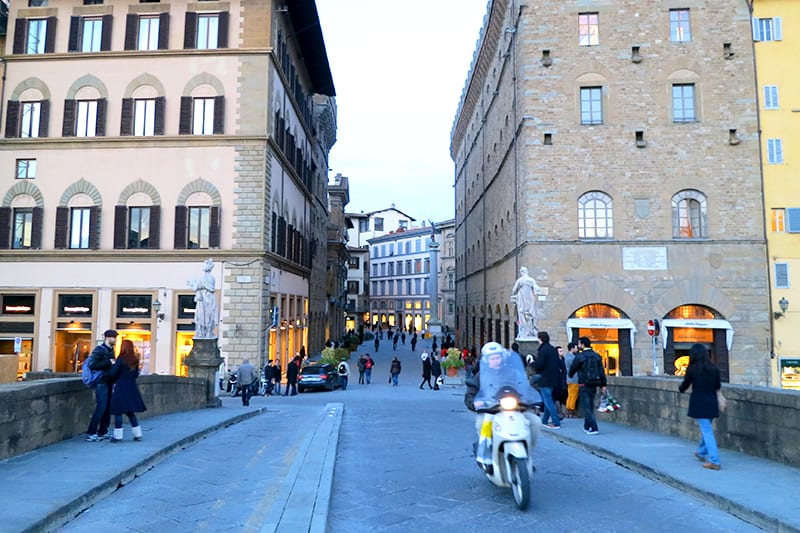 Shopping in Florence - pedestrians walk over a bridge to enter the shopping streets of Florence, as a moped comes the other way towards the camera