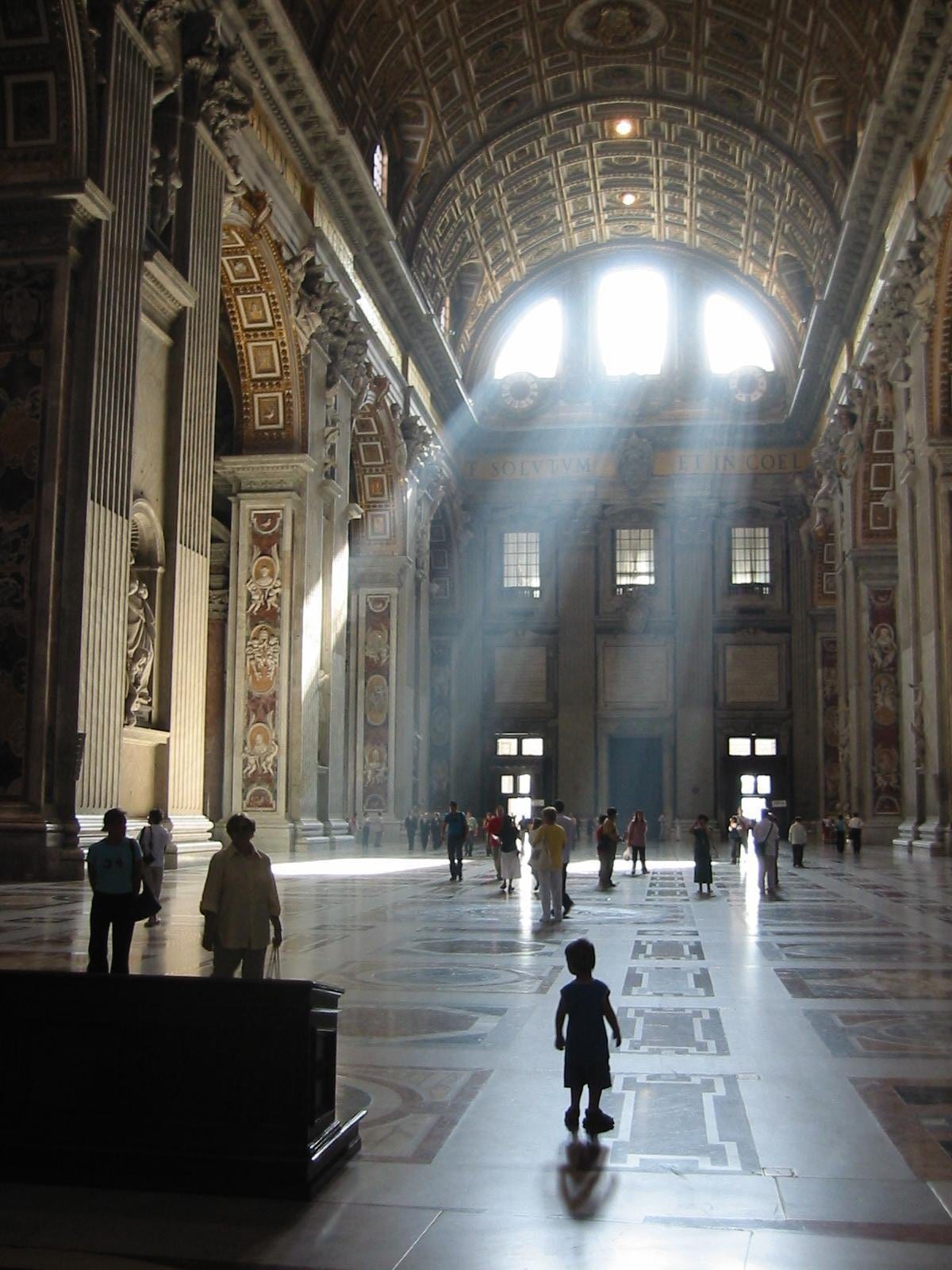 rays of light stream trough high arched windows inside St Peters Basilica, lighting the ornate interior and showing the small silhouettes of people below.