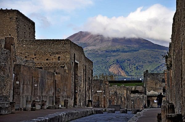 ruined houses on either side of a street in Pompeii, with a smoking mount vesuvius in the background