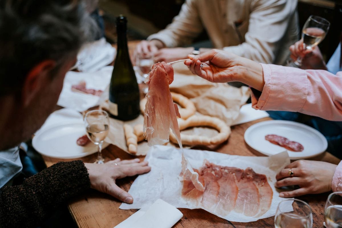 A woman using her fork to select a thin slice of ham off of a plate at a table with others.