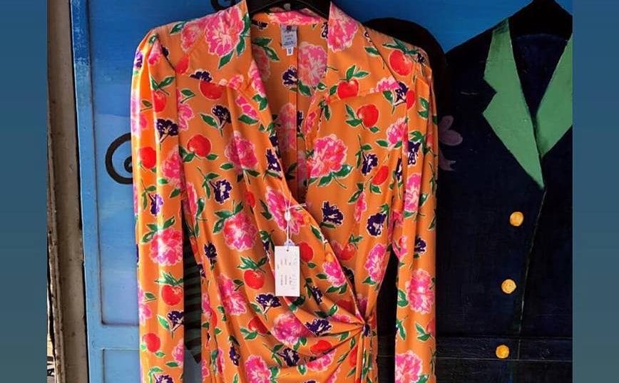 A colourful pink, orange, green floral dress hangs next to a black jacket with green collar, against a blue background at the Mercato-Vintage-Ecosolidale - a vintage market in rome