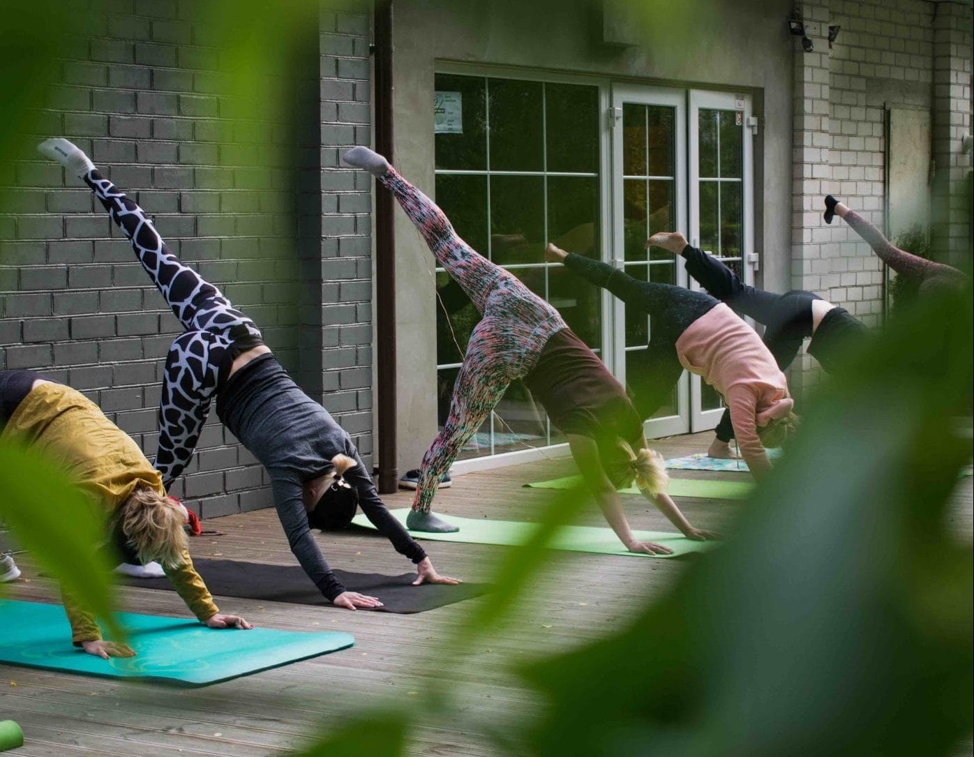 A line of 6 people doing a synchronised yoga pose on mats on decking in a garden outside a building with french windows