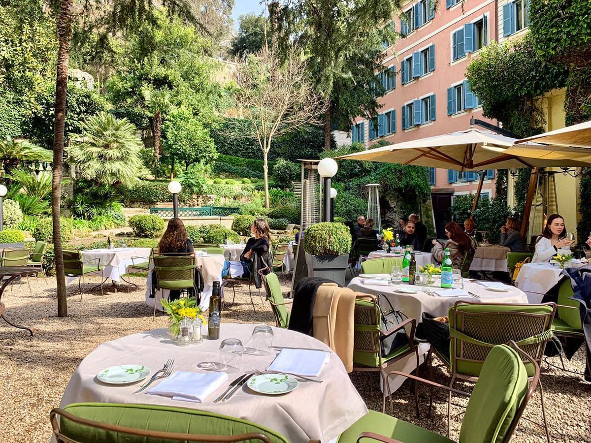 an outside garden or patio area in a hotel, with views of lush green gardens. Decorated with tables and chairs in natural colours, for a relaxed area to dine.