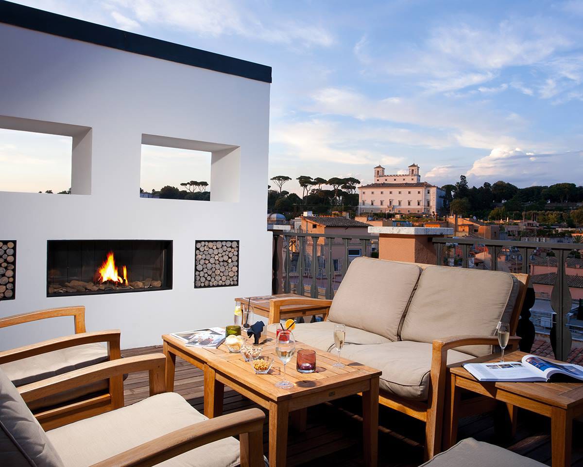 A modern hipster rooftop scene, with faux wall including fireplace and 4 reclining chairs around a coffee table filled with cocktails, and a city view in the distance