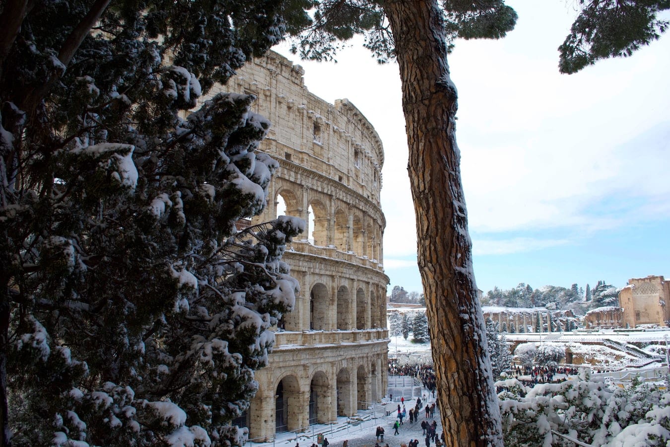 a scenic shot of the the outside of the walls of the colosseum in rome, with snow on the ground and people walking in the distance 