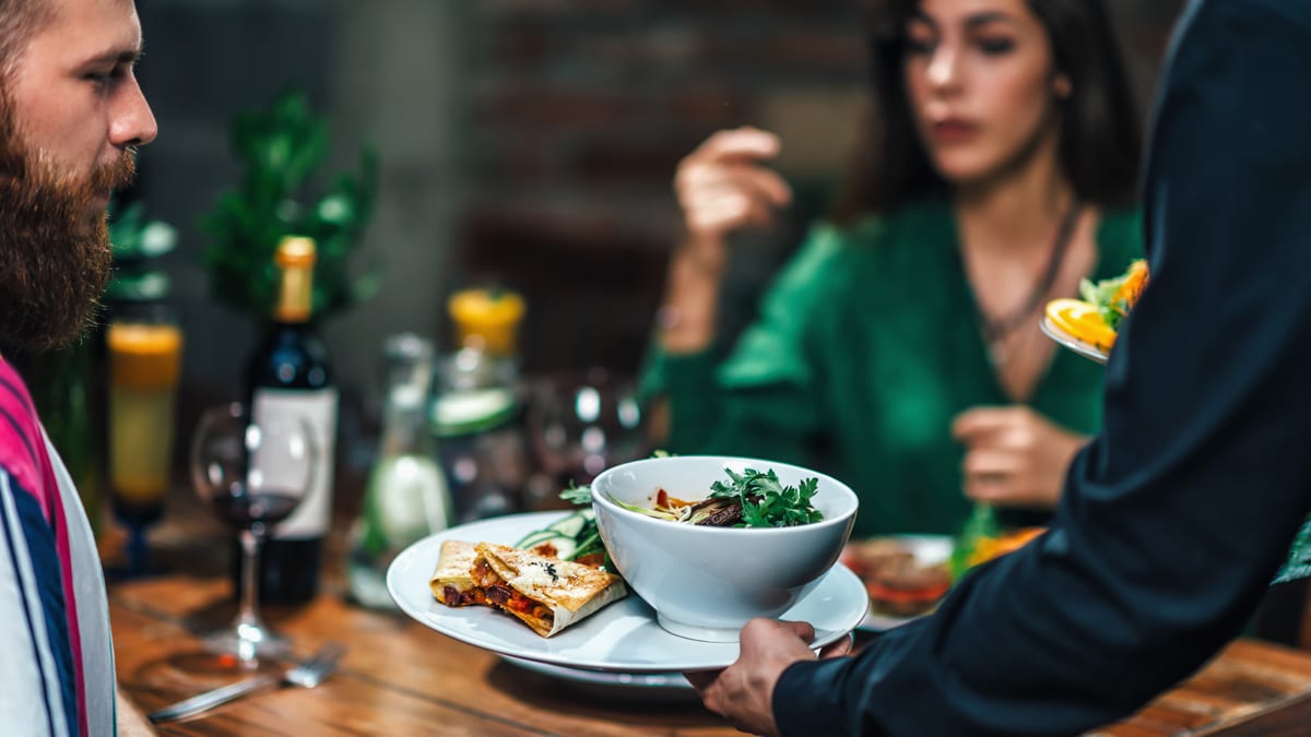 A couple sit either side of a table, set for dinner with wine , as a waiter passes a plate with pasta and flatbreads to them.