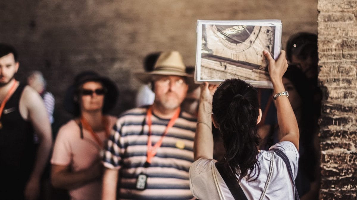 A tour guide with her back to the camera, holds up a picture and shows it to a small group of tourists on the Colosseum guided tour
