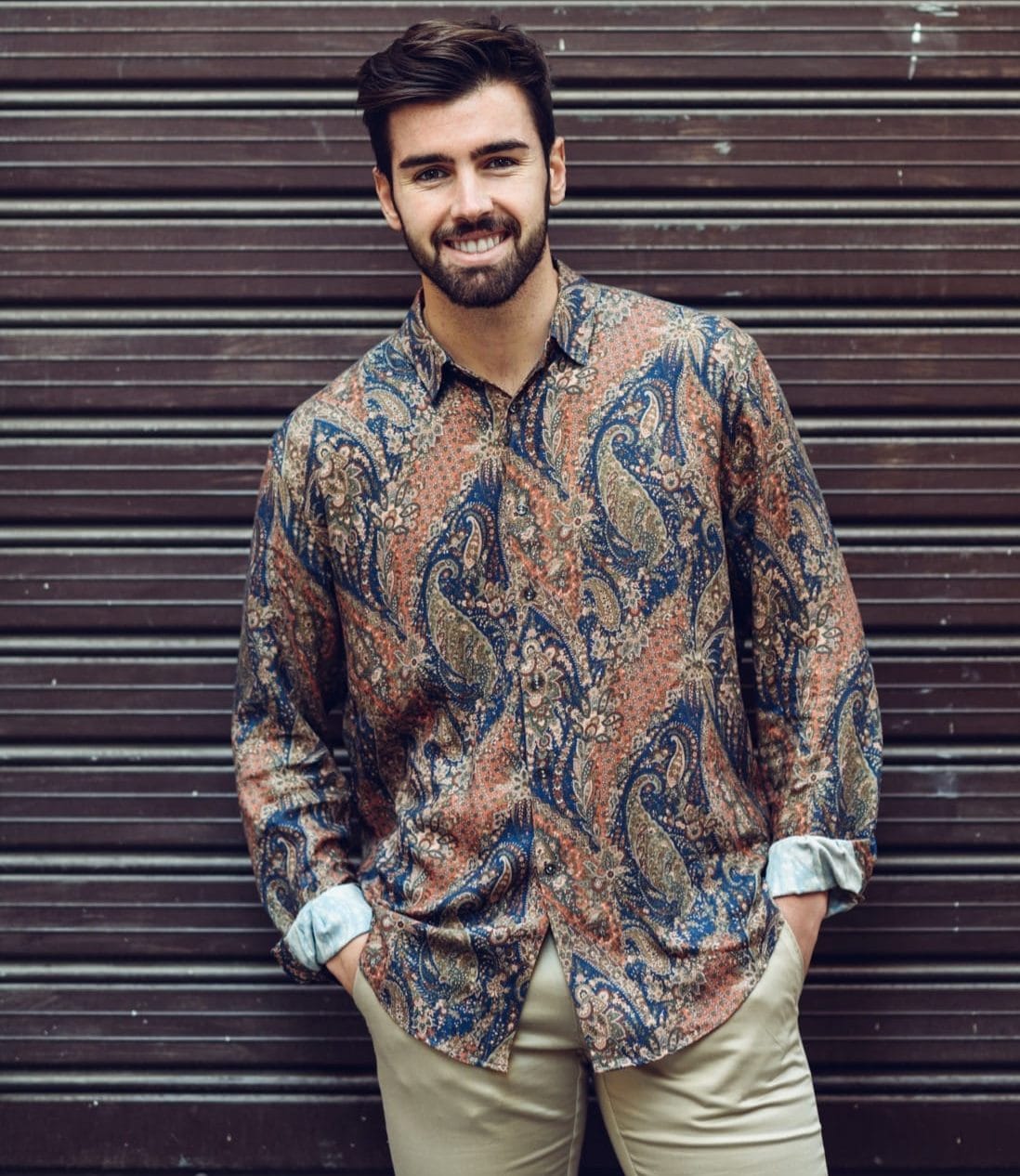 an italian man with a beard stands in a casual stance wearing a patterned shirt, in blue and muted orange tones, and taupe jeans