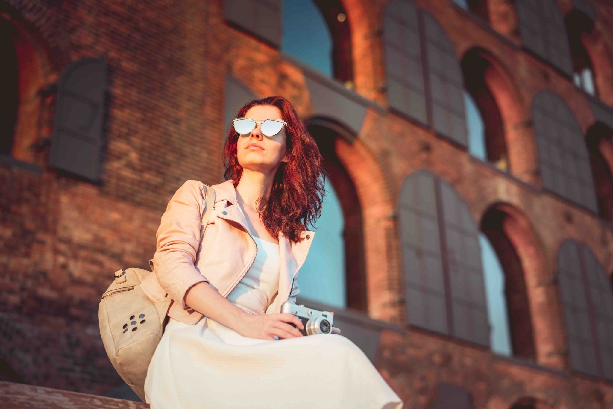 a young women in a seated pose in front of a building with arched windows, with an old -fashioned camera on her lap