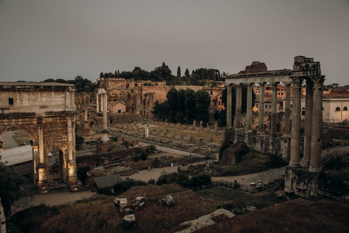 A view of the Roman Forum at dusk, featuring the towering remains of columns and arches. The image highlights the Temple of Saturn on the left, with crumbled stone structures and pathways scattered throughout the ancient ruins.