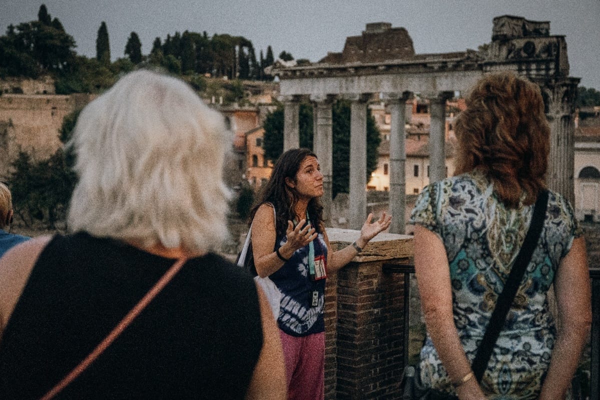 A tour guide explaining Roman history to a group, with ruins in the background at twilight.