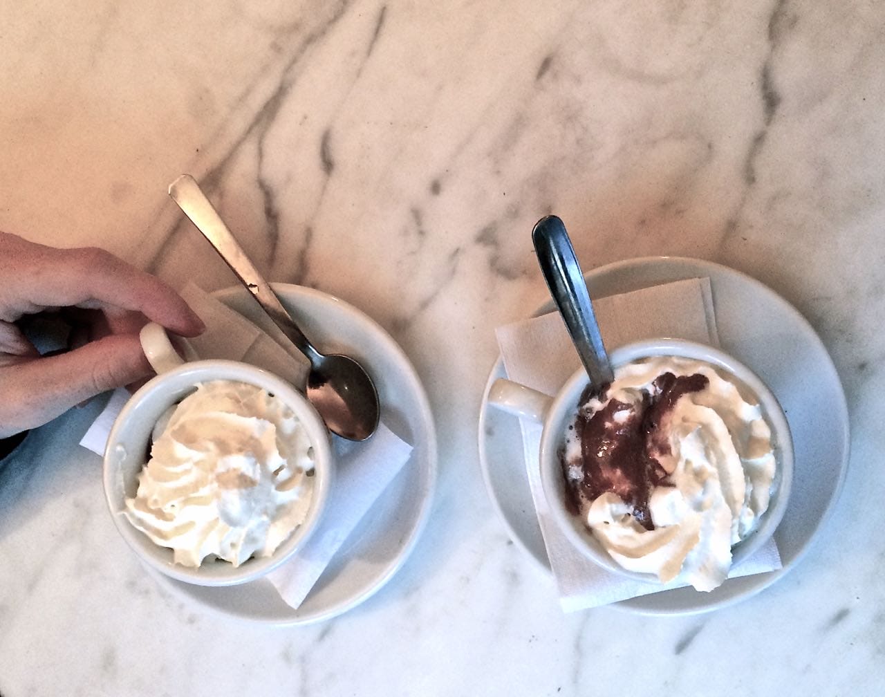 an overhead view of 2 cups of hot chocolate with cream the top, with a saucer and spoon, sitting on a countertop