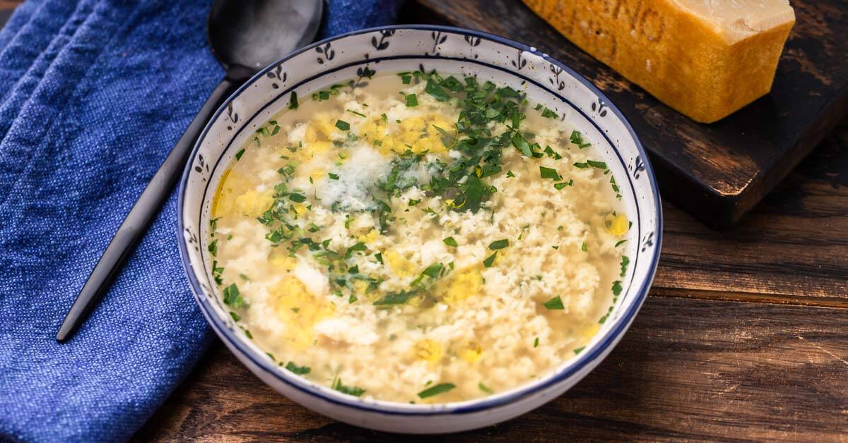 A bowl of Italian Stracciatella soup, in a white and blue bowl, next to a blue cloth and a block of parmesan cheese.