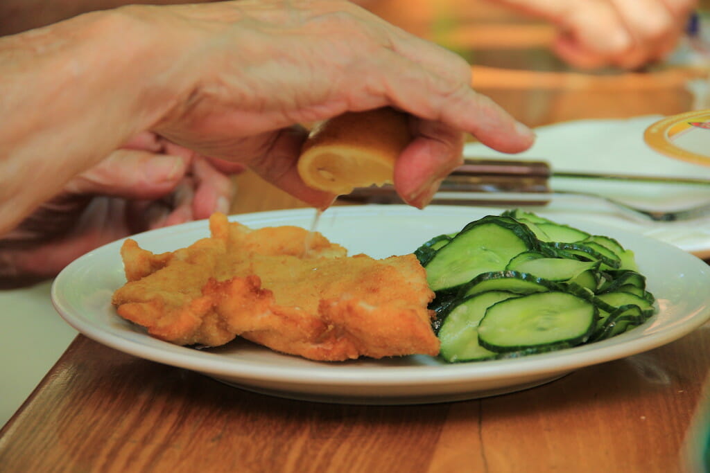 Diner squeezing lemon onto their cotoletta all milanese, with cucumbers on the side of the plate