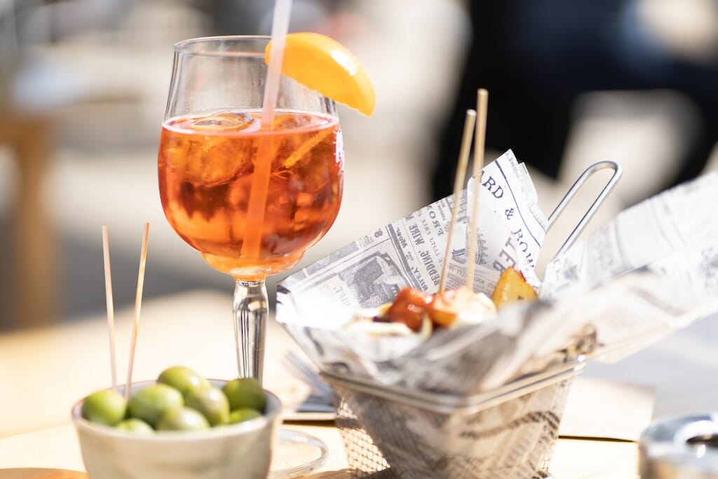 Aperol spritz in a wine glass with lemon along with olives and fried potatoes served outside at a restaurant