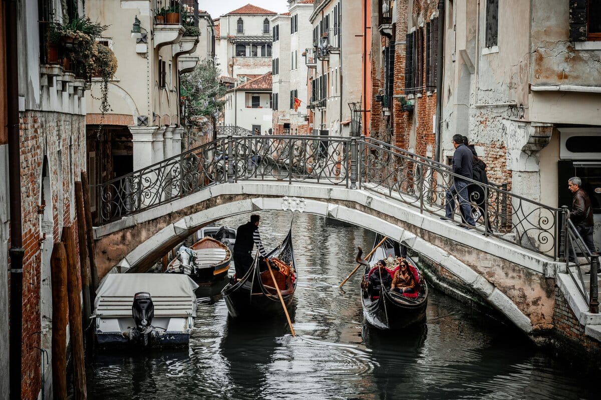 two gondolas in Venice floating under a bridge with people crossing over on top.