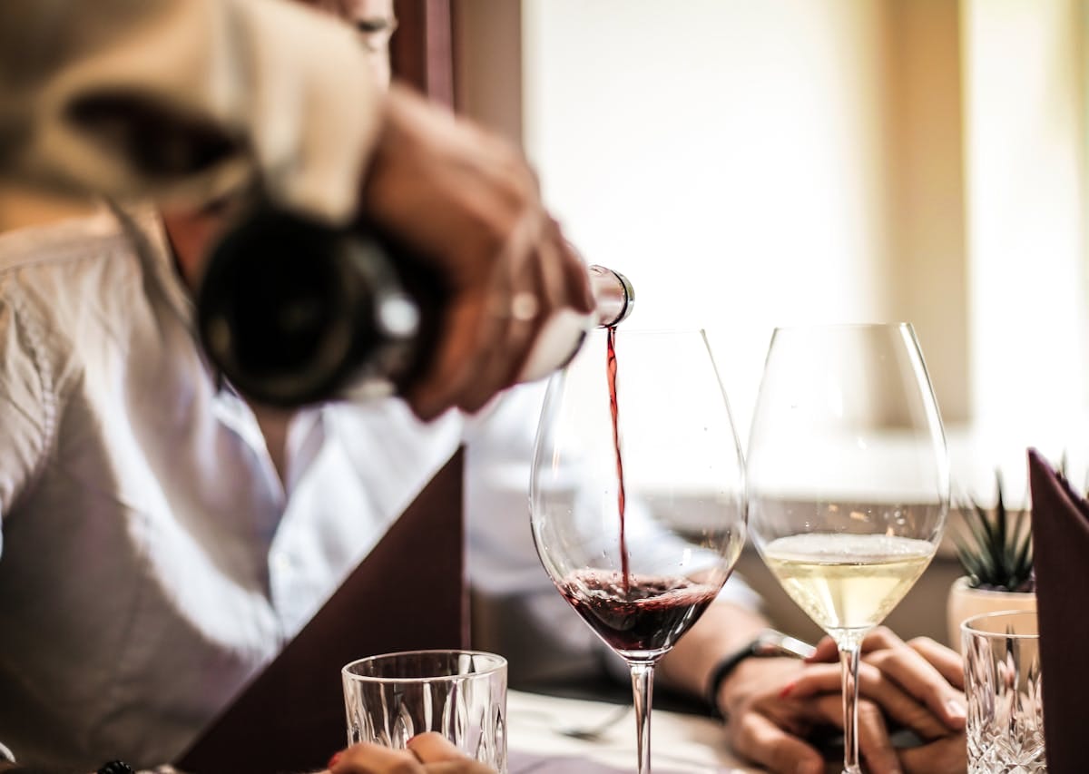 Man pouring red wine into a glass right next to another glass of white wine.