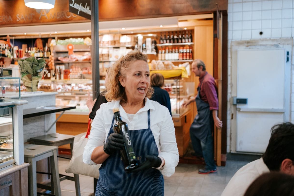 A woman showing a bottle of extra virgin olive oil to a group of people in a food market.
