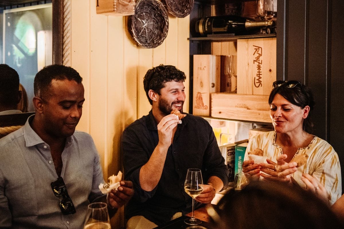 Three people smiling and laughing as they eat food and drink wine.