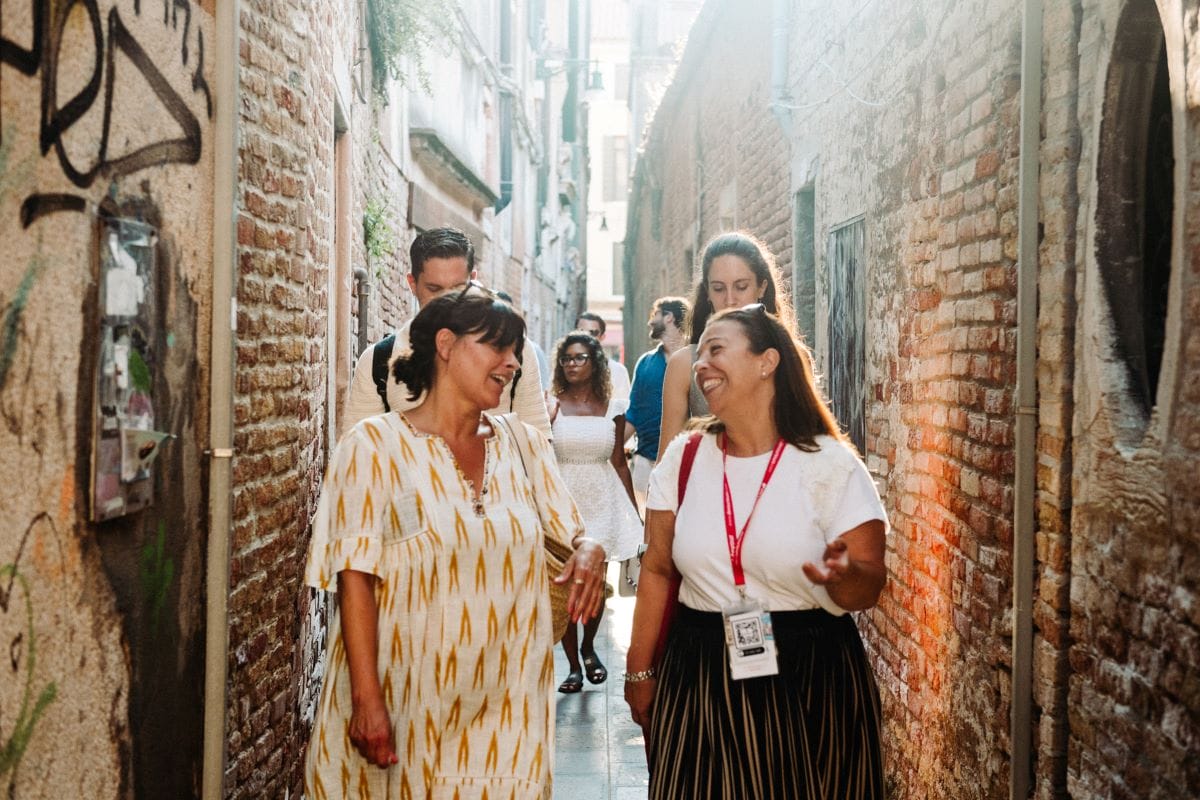 Two women smiling at each other with others following behind them in an alley.