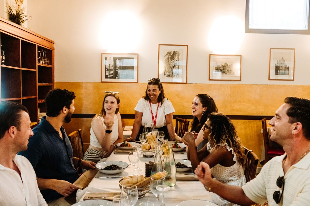 Various people sitting around a large table with plates of spaghett in a black squid ink sauce.