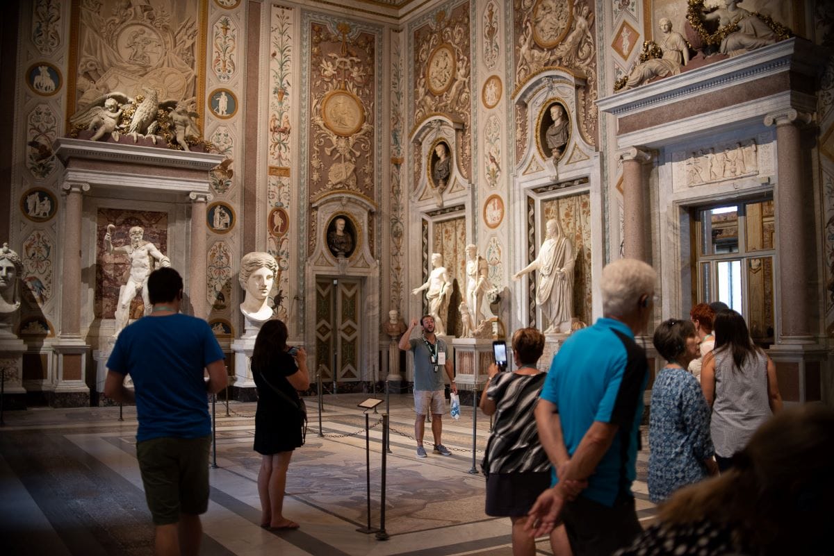 Various people mulling about a large ornate room with statues in the Borghese Gallery.
