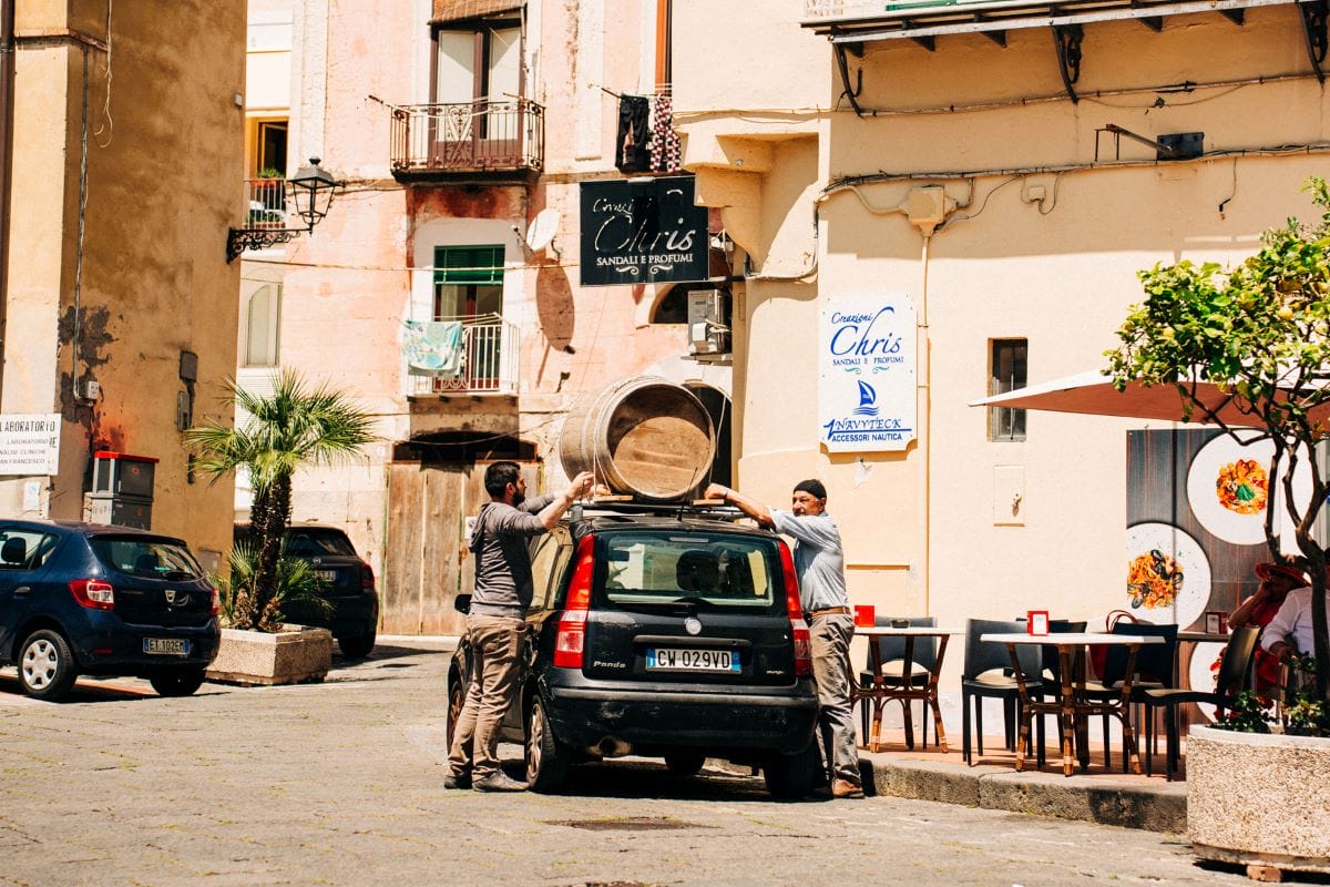 Two men tying a wooden wine barrel to the top of a small car in Italy.