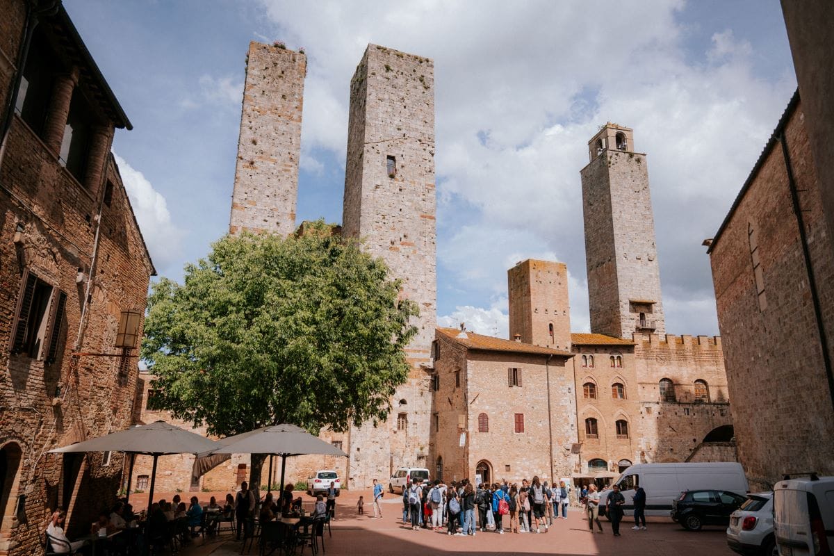 A crowd sits at outdoor tables under umbrellas in Piazza della Cisterna, San Gimignano, Italy, surrounded by medieval stone towers.