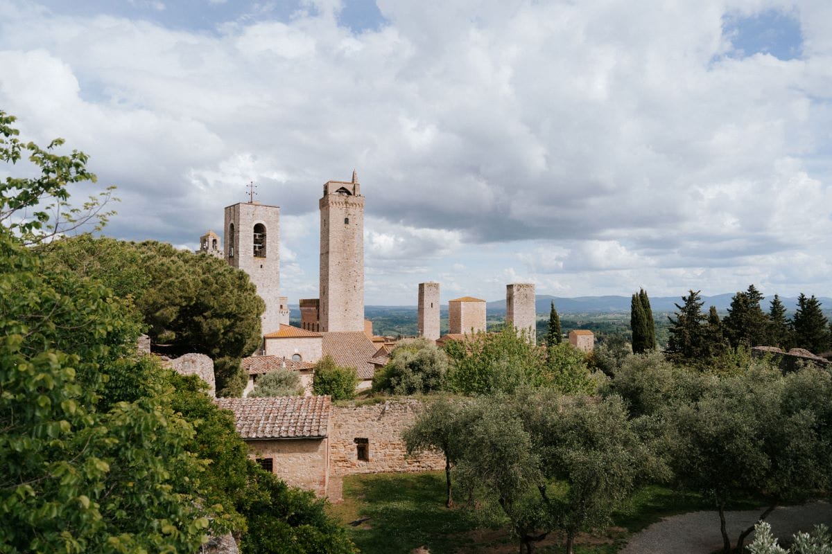 Stone towers stand above green trees and old buildings in San Gimignano, Italy, with a partly cloudy sky overhead.