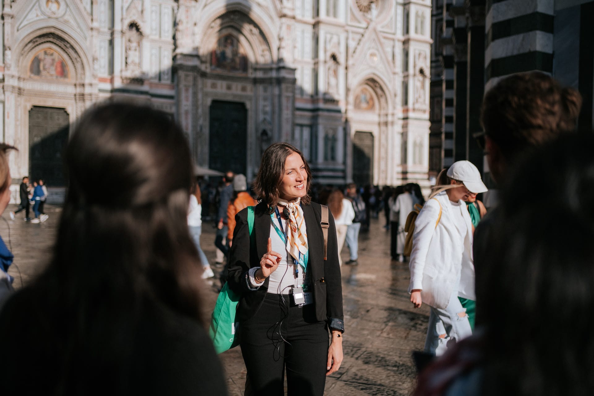 A tour guide speaks to a group of tourists in front of the Florence Cathedral, gesturing with one hand while smiling.