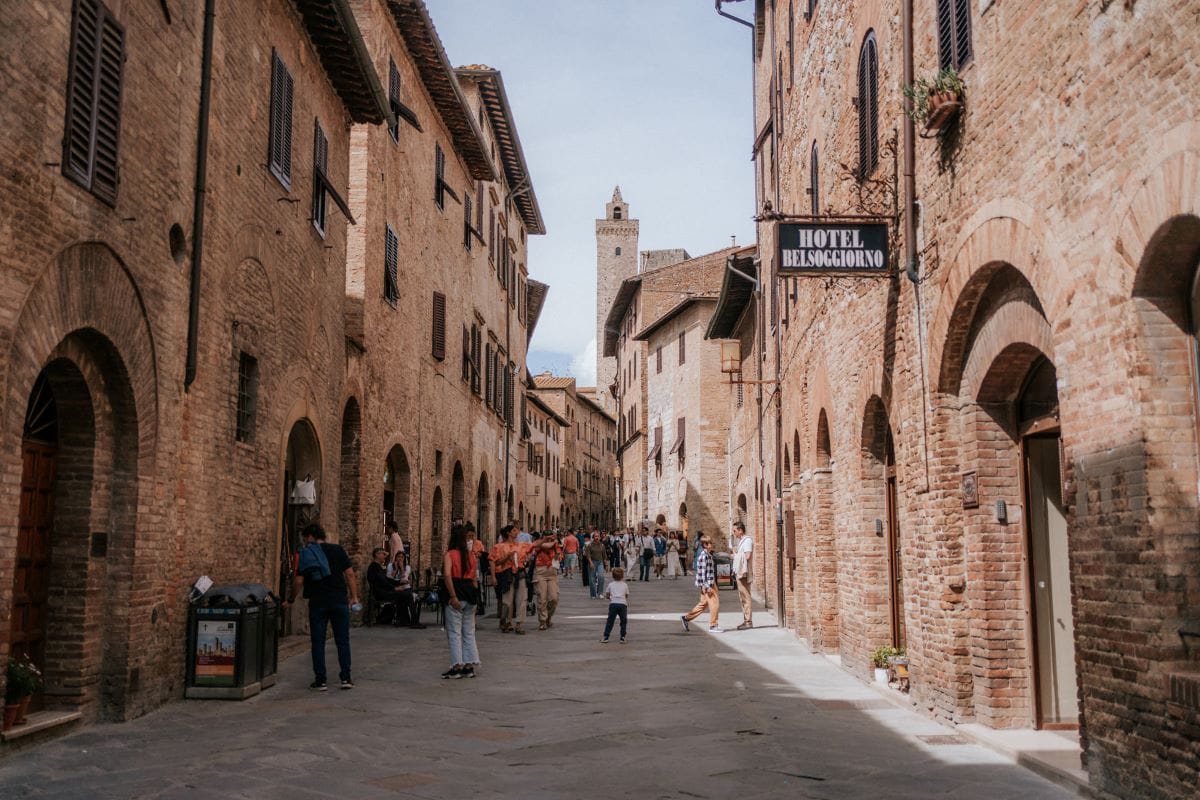 Pedestrians stroll along Via San Matteo in San Gimignano, Italy, passing Hotel Bel Soggiorno’s sign and medieval tower.
