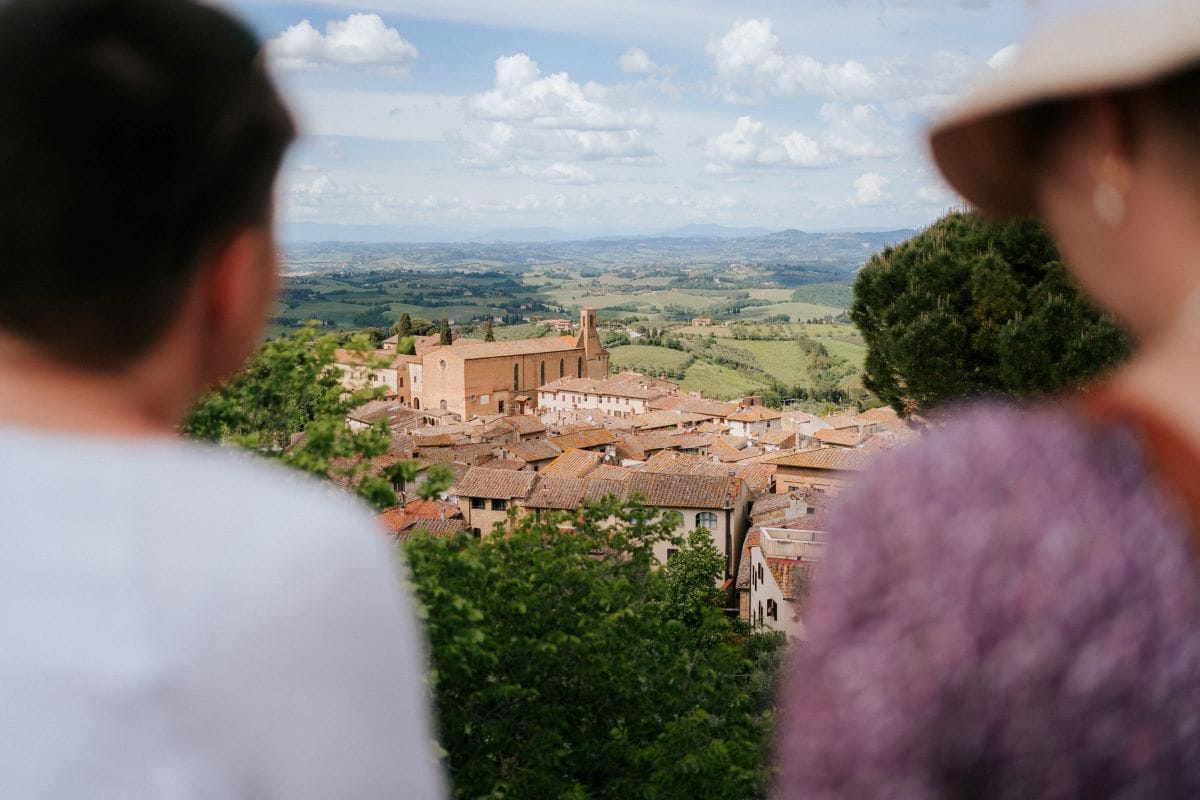 Two people stand and look at the hilltop town of Piazza San Pietro, Tuscany, Italy, with red rooftops and green hills.