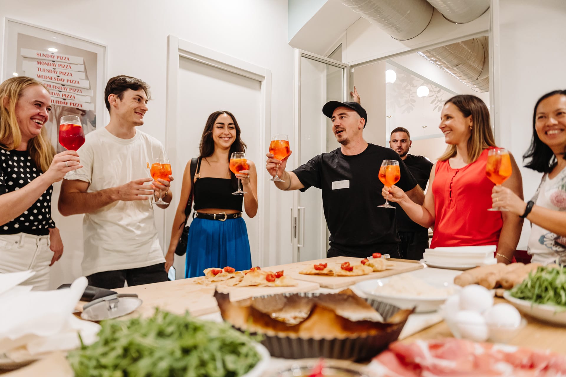 A group of people raising glasses filled with orange cocktails in a cheerful toast around a kitchen island topped with fresh pizza ingredients.