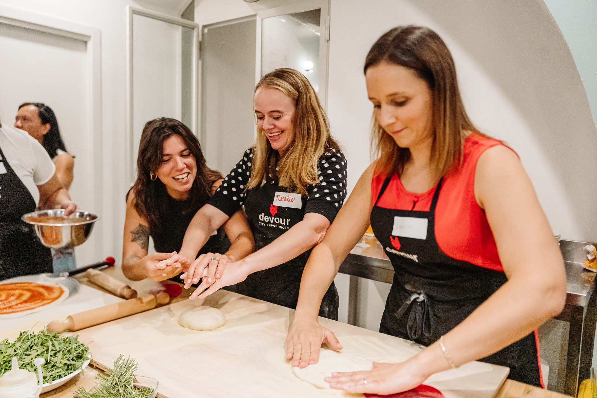 A group of 3 women wearing aprons stretching out pizza dough, on a kitchen counter.