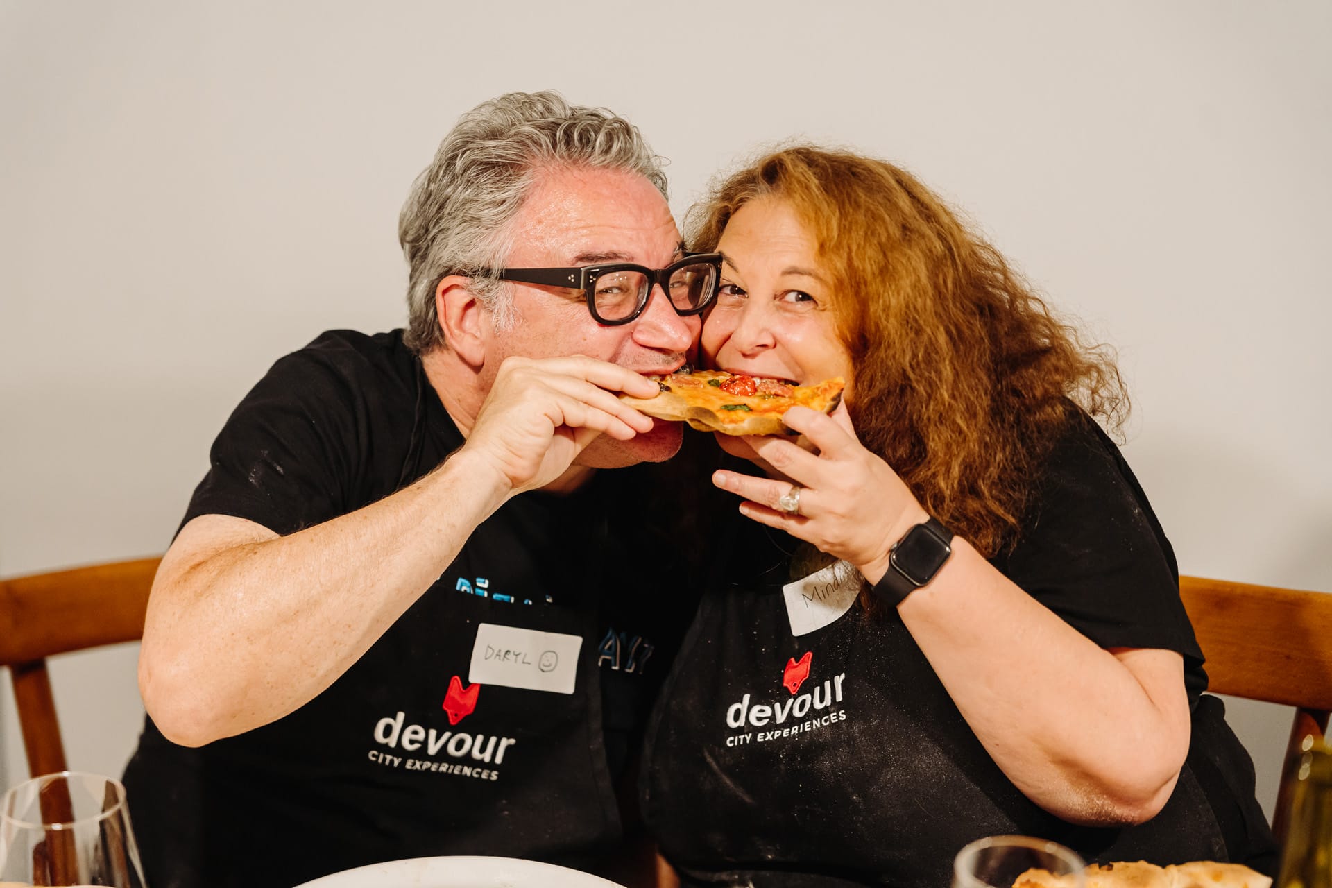 A man and a woman joyfully sharing a slice of pizza, both wearing aprons with “Devour City Experiences” logos, clearly enjoying the moment.