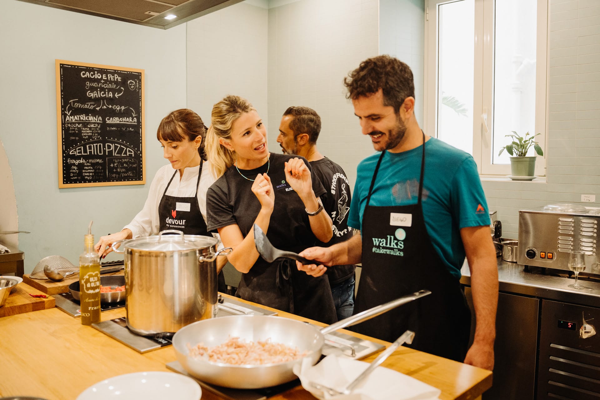 Four people in aprons enjoy a cooking class, chatting and preparing food around a kitchen counter with a chalkboard menu in the background.