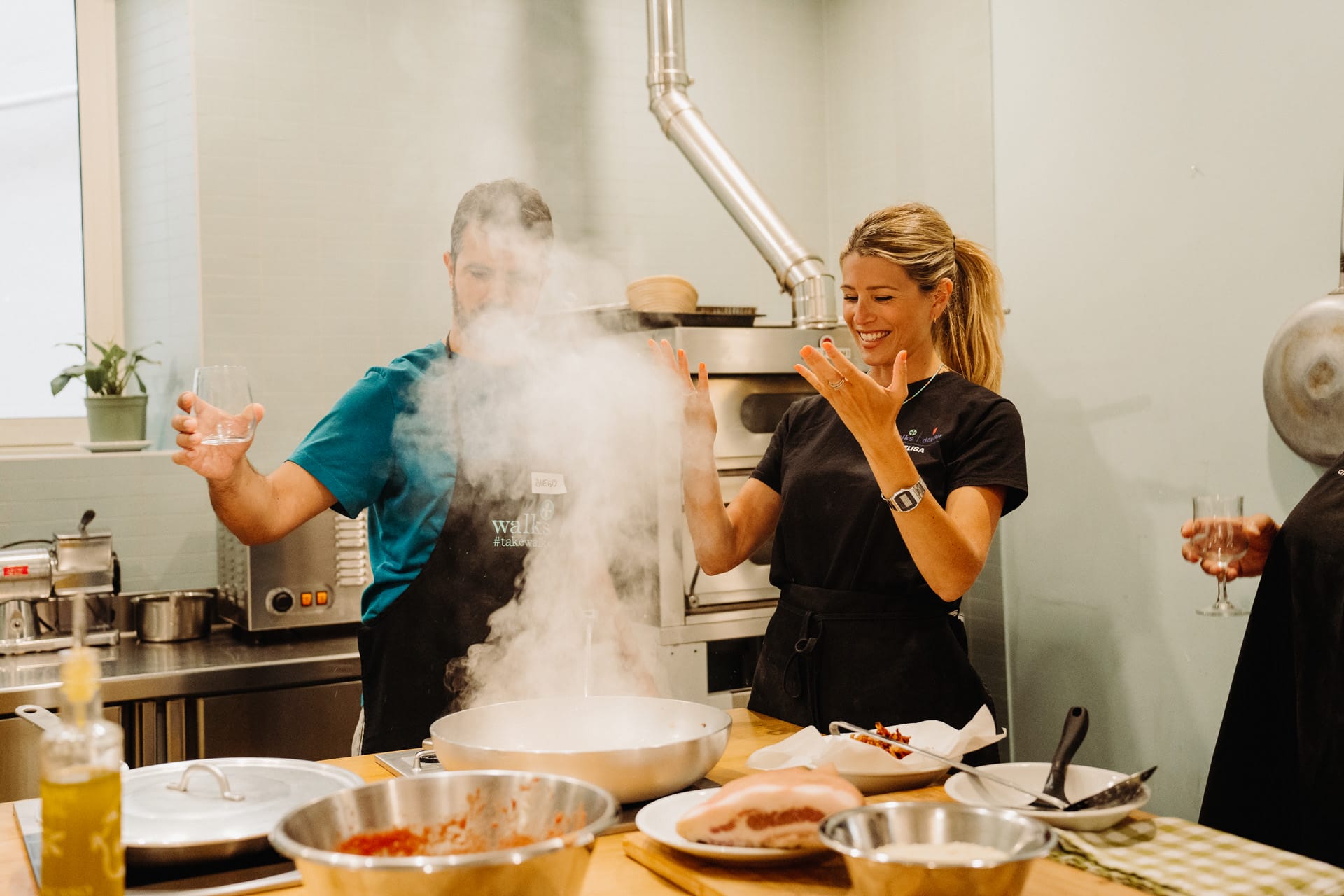 Two cooking participants reacting with joy and surprise as steam rises from a hot pan in a Roman kitchen.
