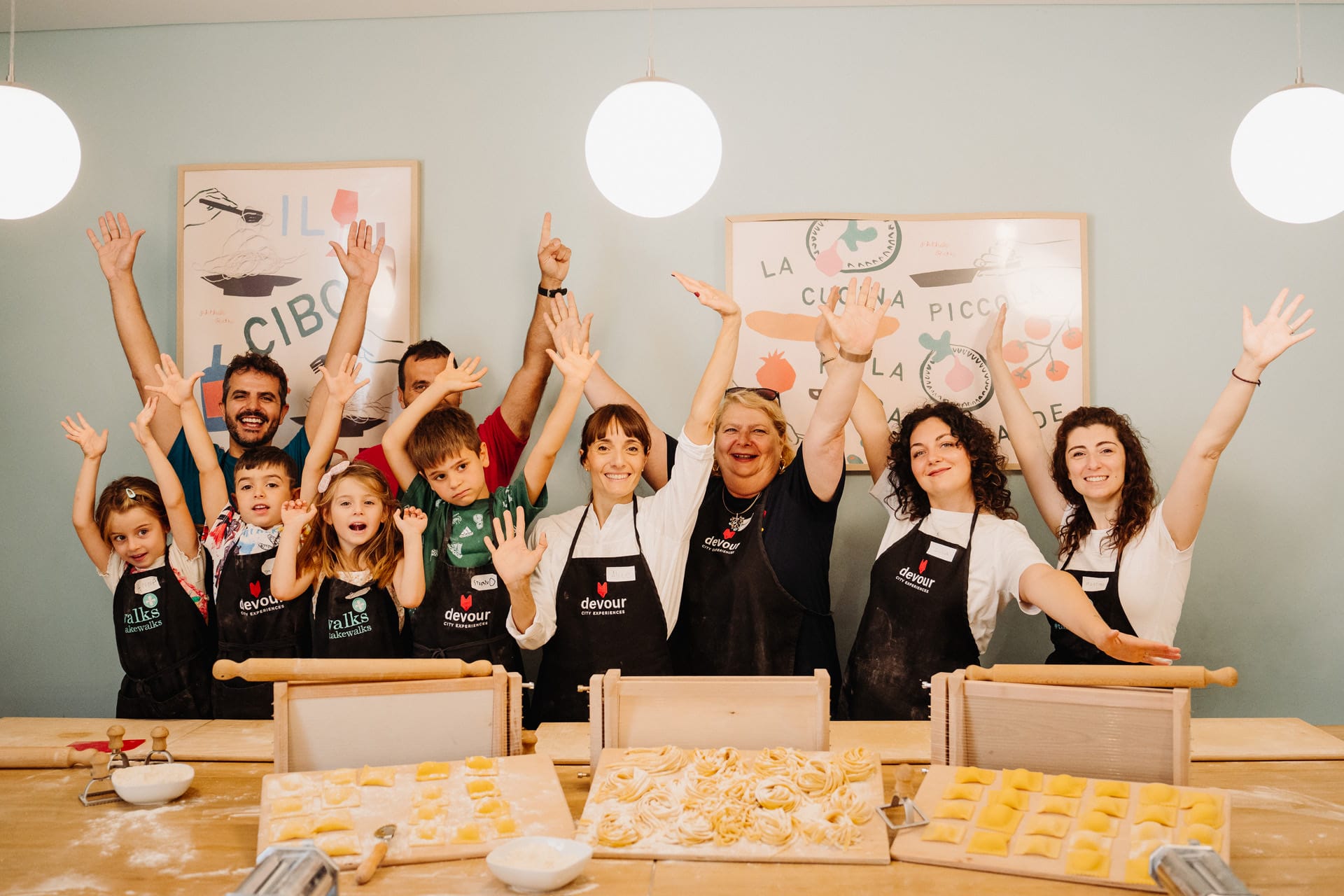 A joyful group of cooking attendees, both kids and adults, celebrating around a table filled with handmade pasta shapes.