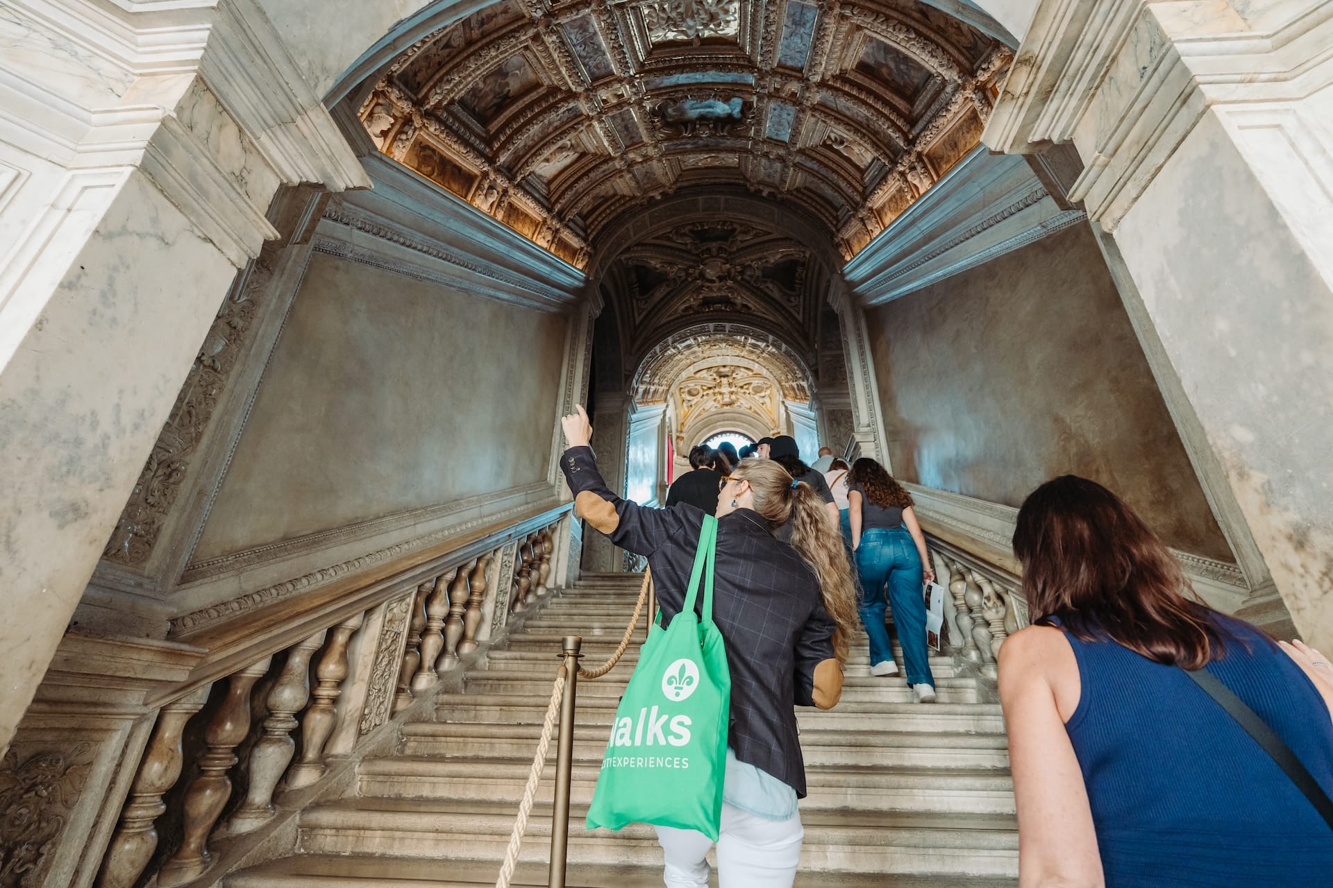 A tour group ascends an ornate marble staircase beneath a richly decorated ceiling in a historic Venetian palace.