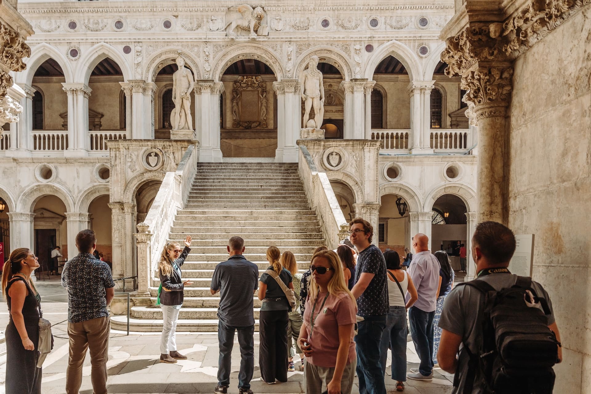 A tour guide speaks to a group of tourists in front of an ornate marble staircase in the courtyard of a historic palace.