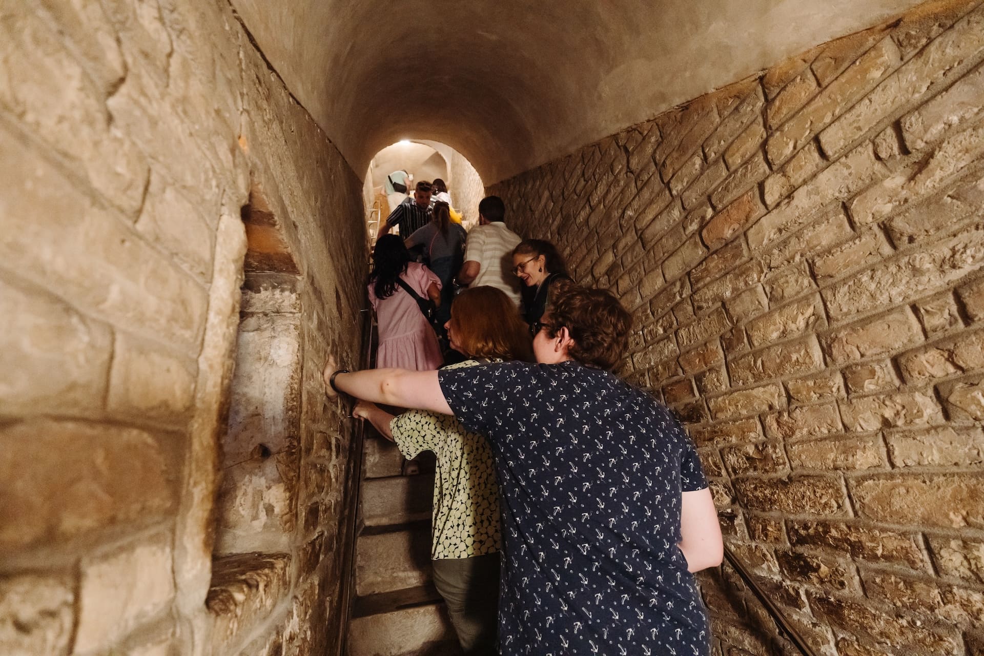 A group of people climb a narrow, dimly lit stone staircase inside a historic tunnel-like corridor.