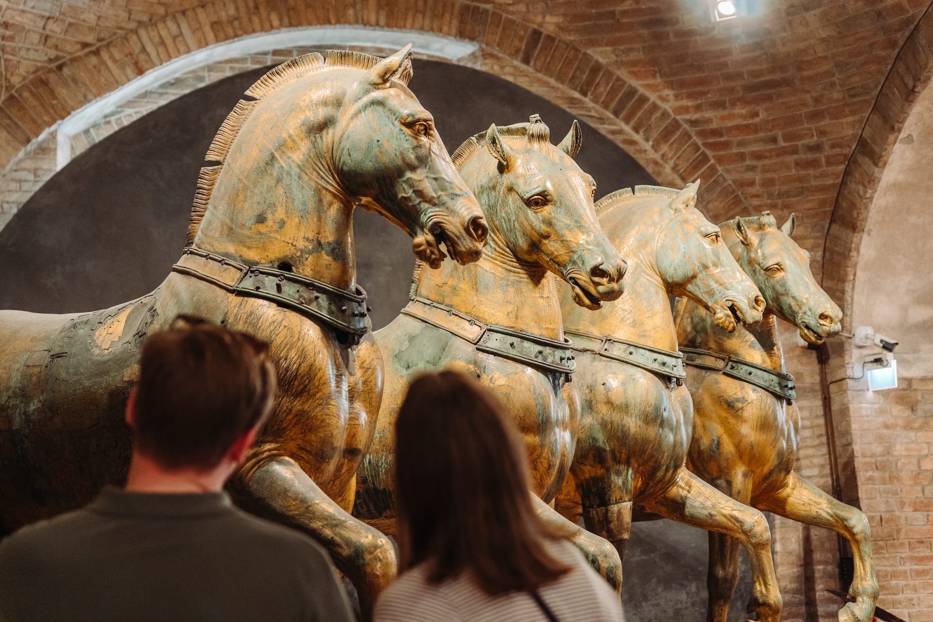Two visitors admire the famous bronze Horses of Saint Mark displayed inside a museum with brick archways.