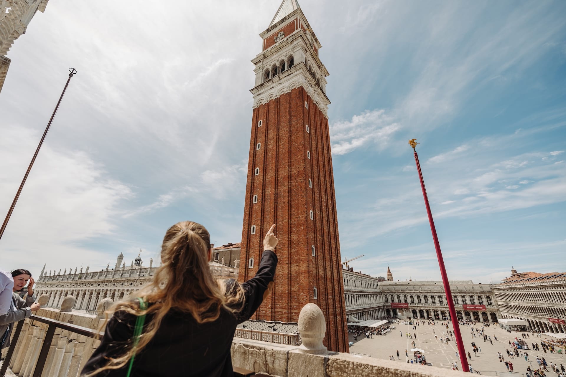 A woman points up at the Campanile di San Marco in Venice, Italy, with a bright blue sky overhead.