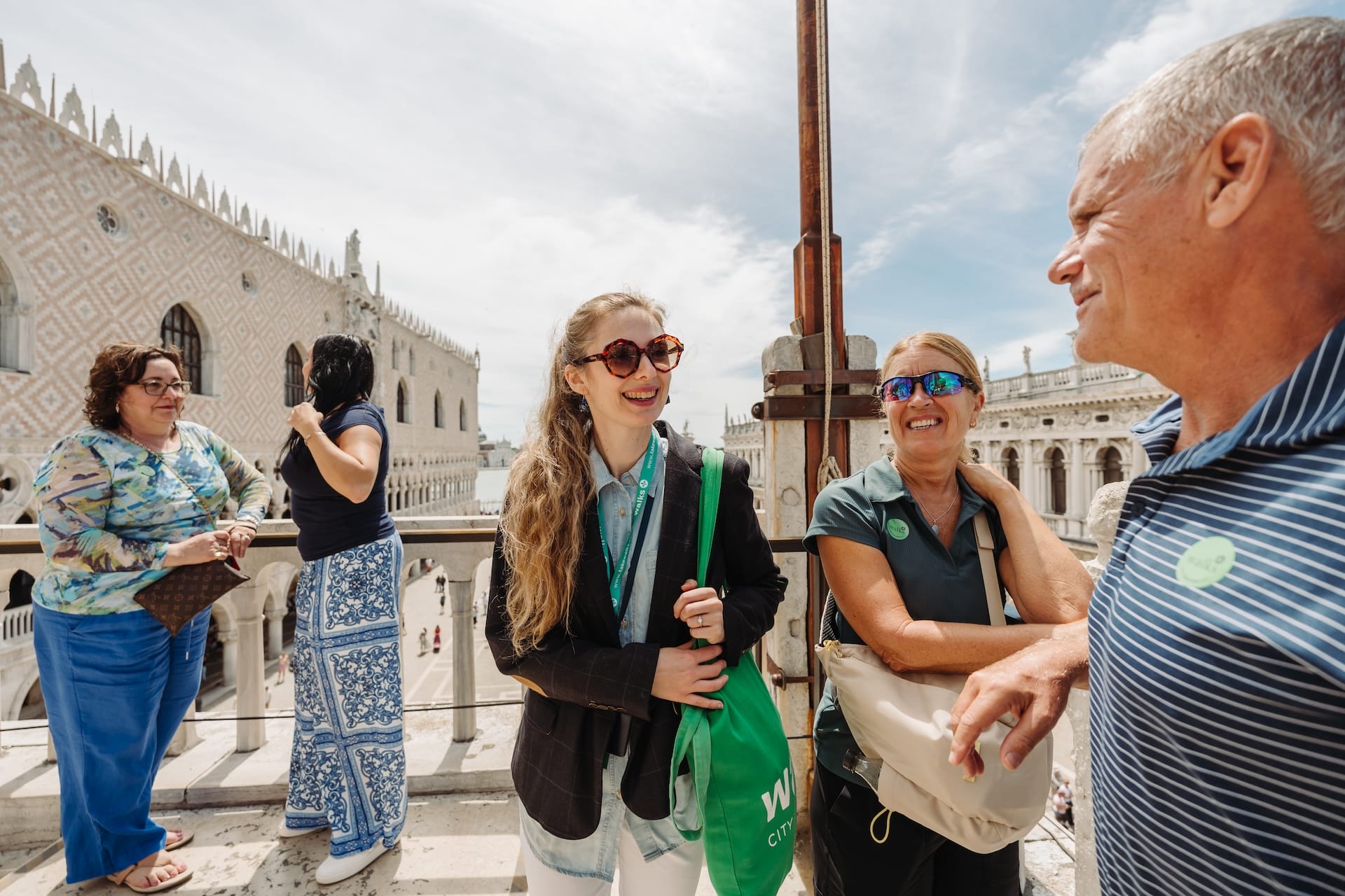 A tour guide is speaking to a couple about the historical sites of Venice during a guided tour. There are 4 visitors ansdone guide standing on a roof terrace of one of the central venice attraction