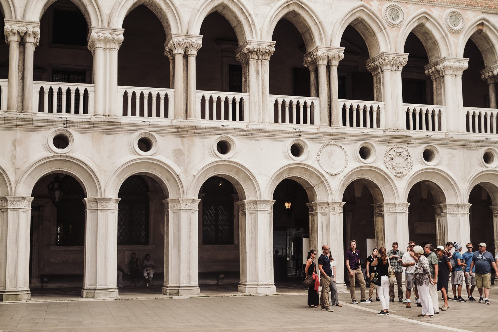 A group of tourists stands in the courtyard of a Renaissance building with arched colonnades and intricate marble details.