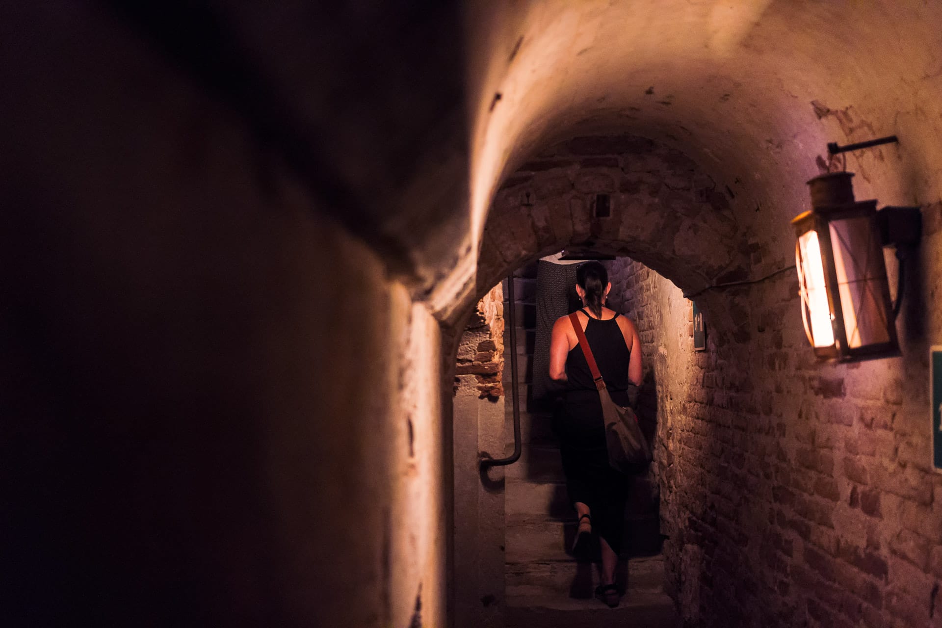 A woman walks through a dimly-lit, narrow stone passage with a lantern hanging on the wall.